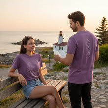 Charger l'image dans la galerie, Couple portant des t-shirts Impressions Gaspésie en coton épais violet avec motifs, assis sur un banc près d'un phare.