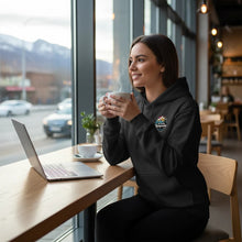 Charger l'image dans la galerie, Femme portant un hoodie noir Impressions Gaspésie avec logo, assise dans un café avec un ordinateur et un café chaud.
