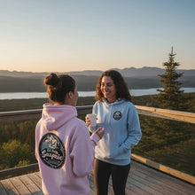 Charger l'image dans la galerie, Deux femmes portant des hoodies Impressions Gaspésie pastel, rose et bleu, sur une terrasse avec vue panoramique.
