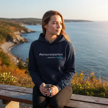 Charger l'image dans la galerie, Femme portant un ouaté à capuche bleu marine Impressions Gaspésie avec inscription #lamourestpartout au bord de la mer
